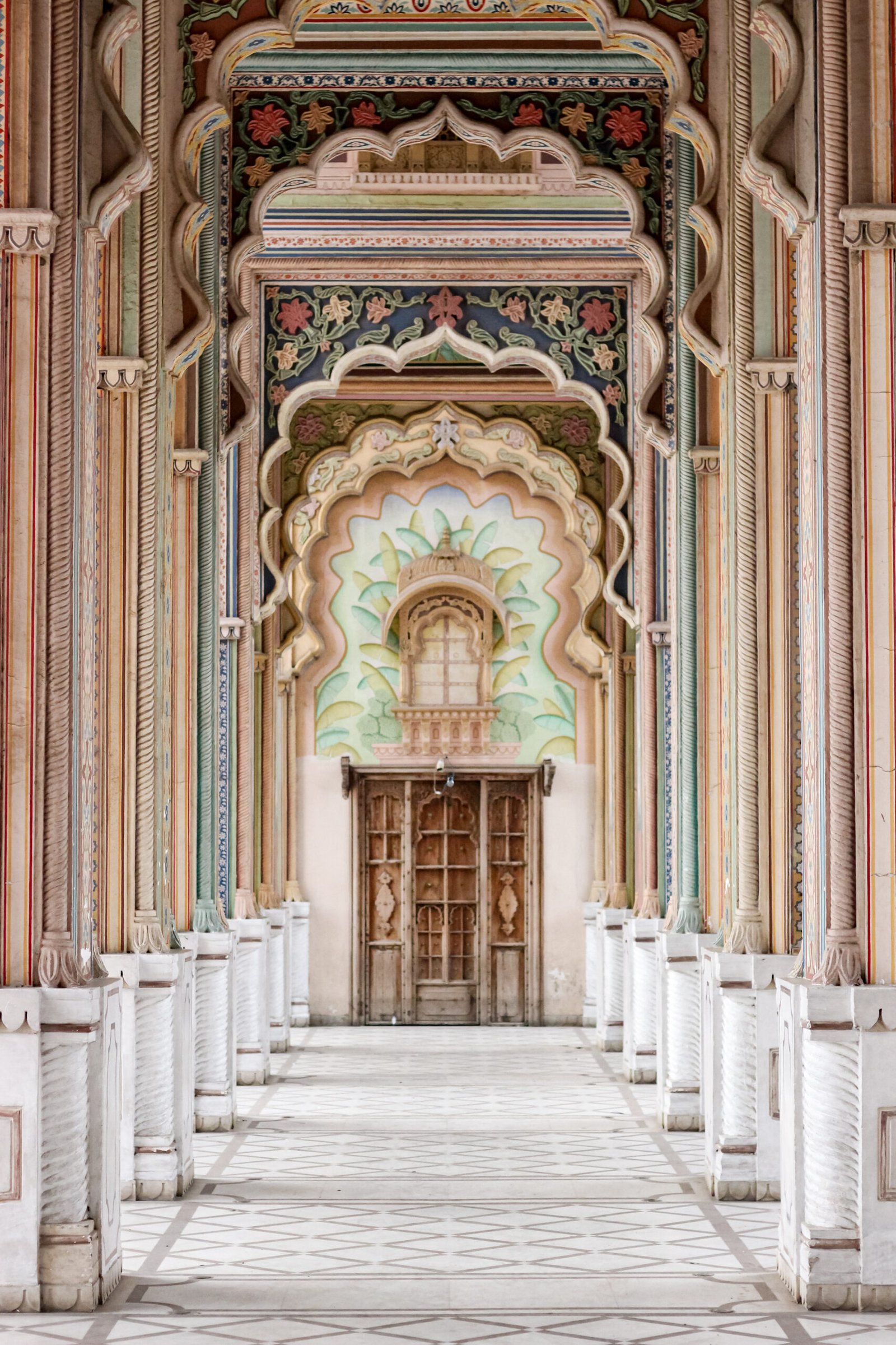 A symmetrical view of a grand, ornately decorated corridor in India featuring intricate floral patterns and multiple layers of colorful, carved arches leading to a wooden door.