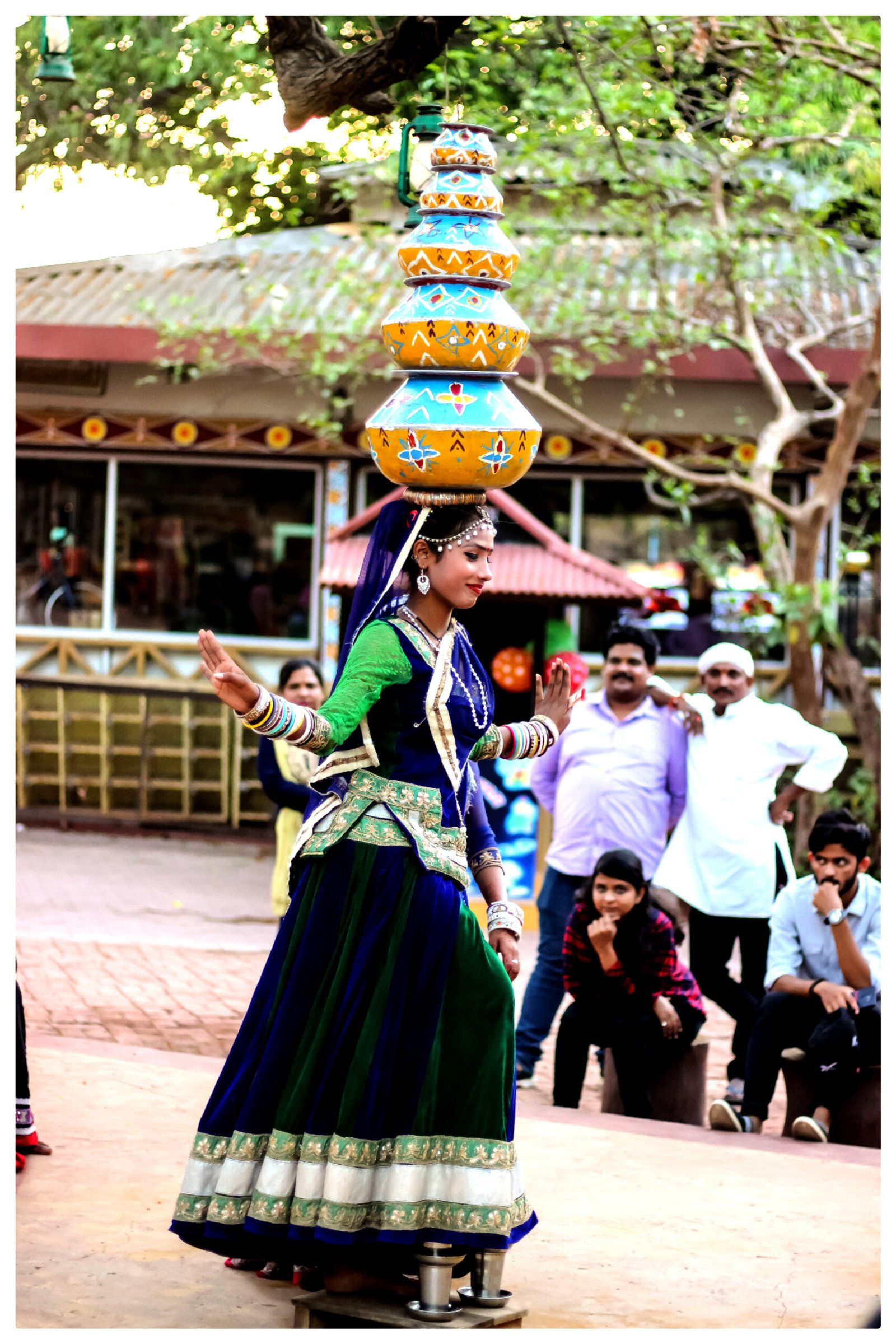 A woman in vibrant blue and green traditional Indian attire performing a folk dance while skillfully balancing several colorful, stacked pots on her head.